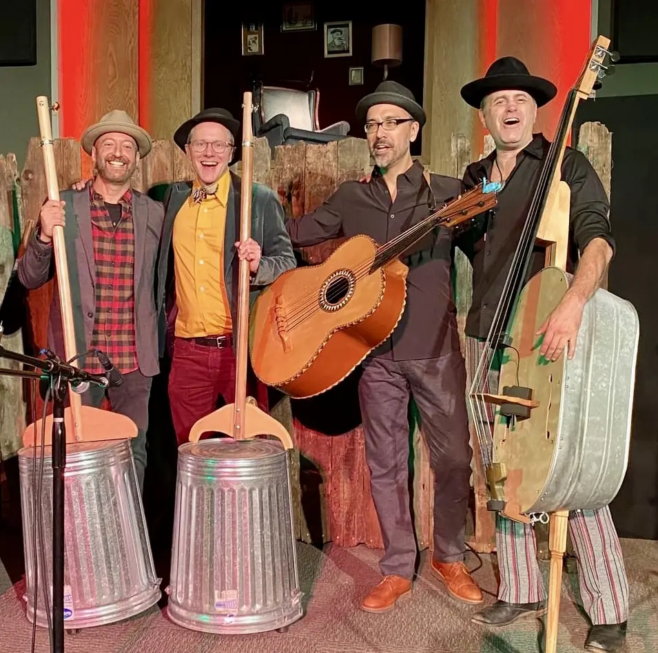 Four grinning musicians in hats on stage: two with trashcan basses, one with a washtub bass, and Jason Krivo Flores holding a guitarrón